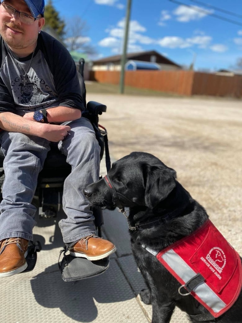 A man sits in a motorized wheelchair. A black dog in a red vest sits in front of him.