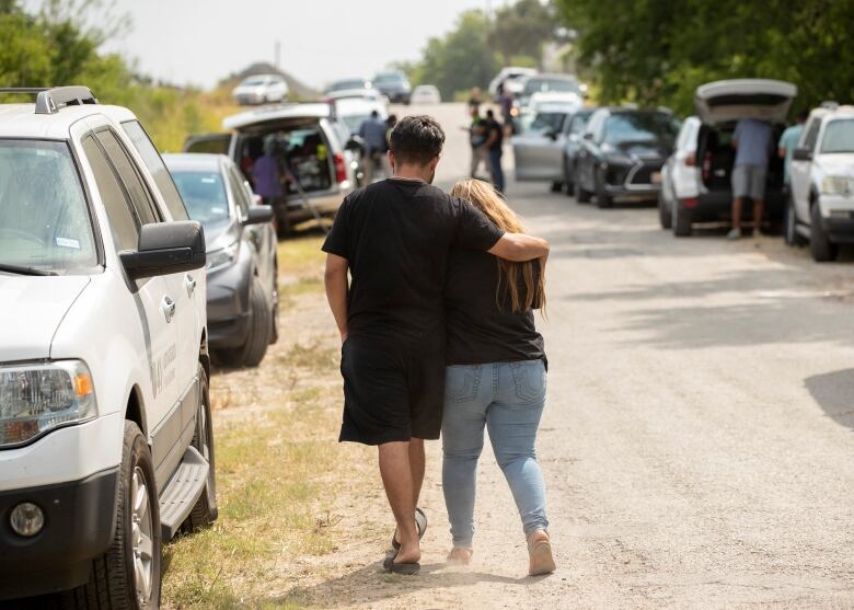 A man and woman walking on a dirt road past several parked cars on either side. The man has his arm around the woman's back. 