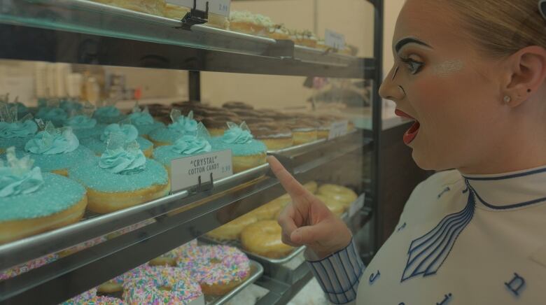 A woman with face paint on points to doughnuts in a display case.