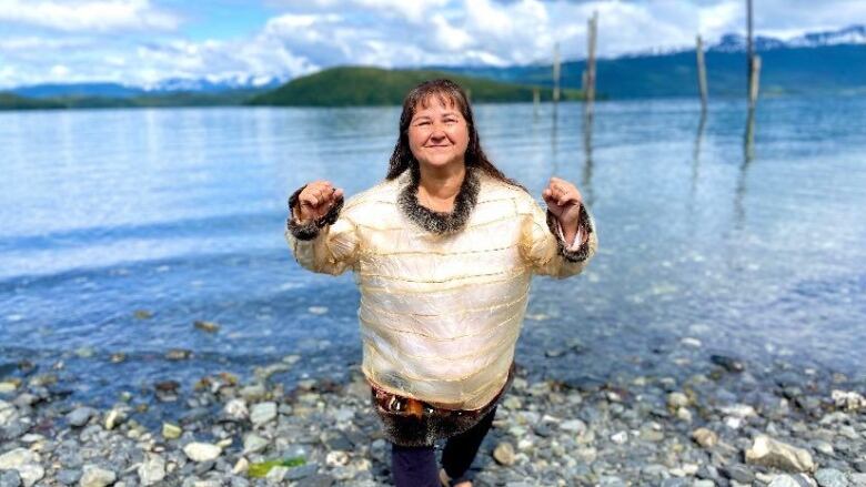 A smiling woman stands on a rocky beach, proudly wearing a transparent parka with fur details.