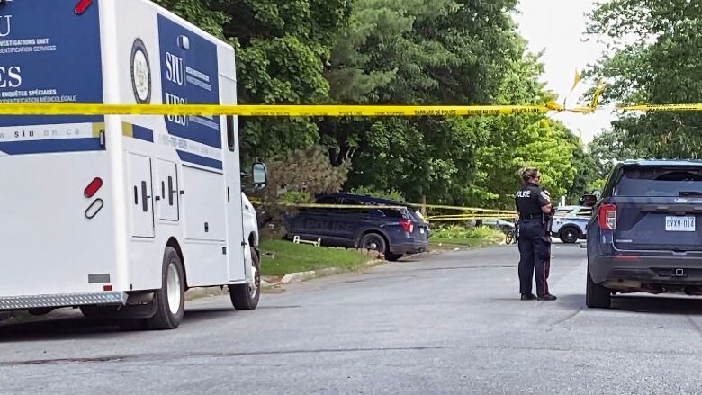 Yellow police tape across a residential street with police and SIU vehicles parked on either side. A female police officer stands near the middle of the road.