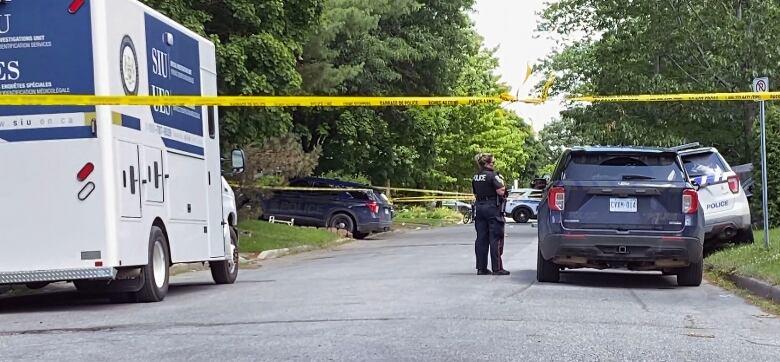 Yellow police tape across a residential street with police and SIU vehicles parked on either side. A female police officer stands near the middle of the road.