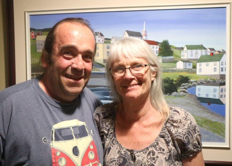 A smiling man and woman pose in front of a painting of a rural Newfoundland scene.