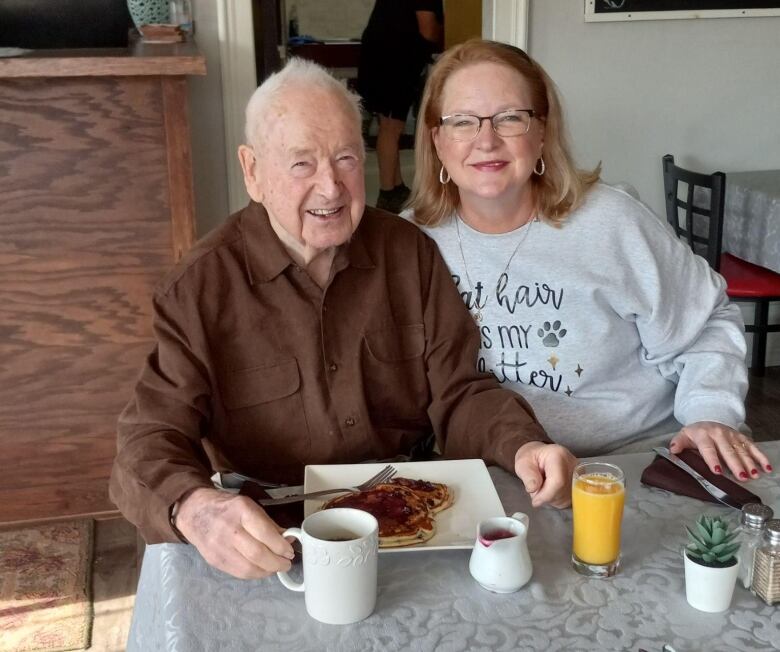 A man and woman look at the camera while sitting at a restaurant table. A plate of breakfast is before the man. 