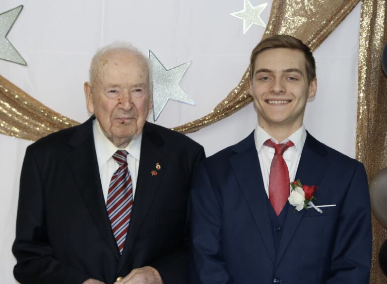 An older man stands next to a smiling teenager, with prom decorations in the background. 