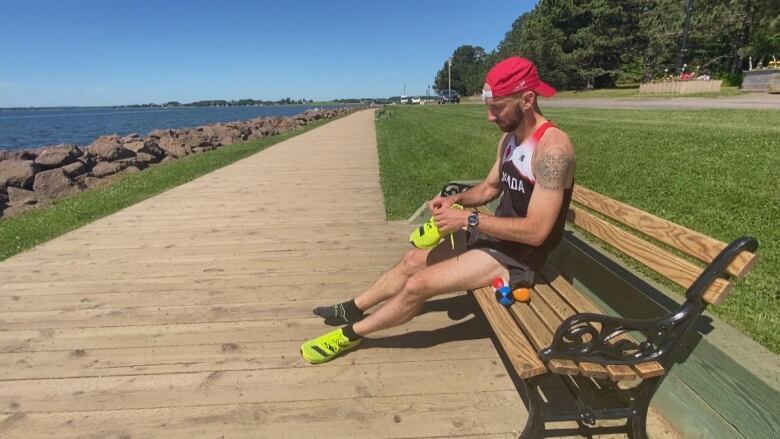 Man in baseball hat, tank top and shorts sitting on a park bench next to a trail, putting on a sneaker.