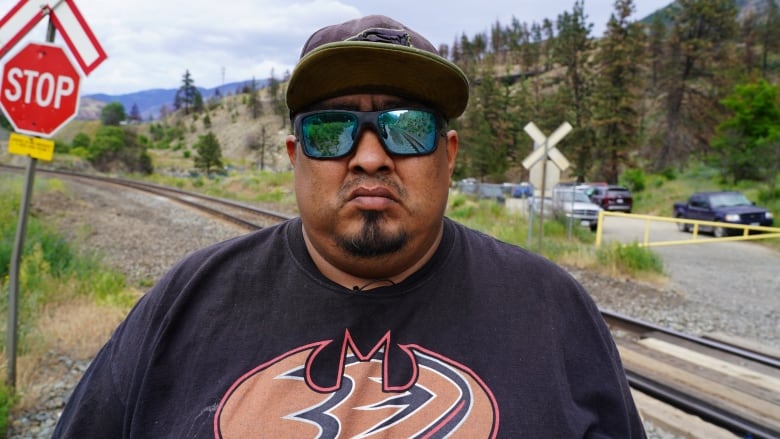 A man wearing sunglasses stands in front of railway tracks.