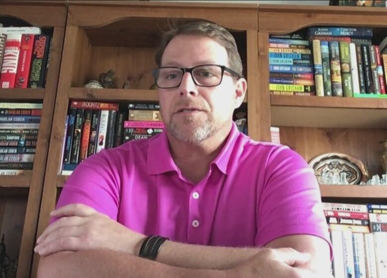 A person wearing a pink shirt and glasses sits in front of a book shelf with their arms crossed.