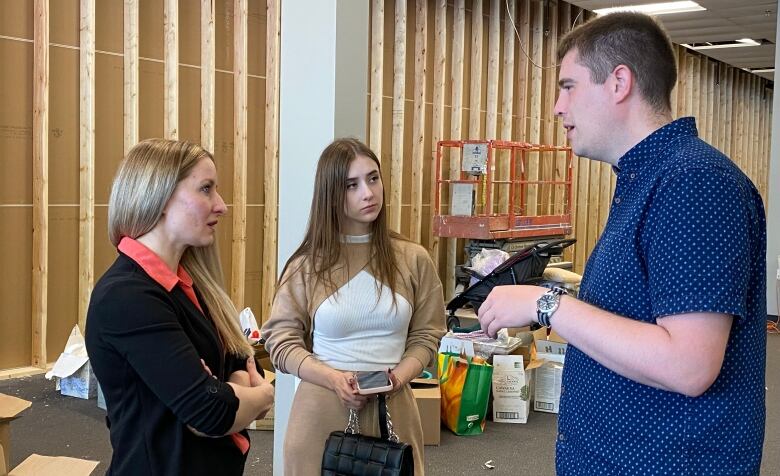 A man talks with two women in an open warehouse space, with bags and boxes in the background.