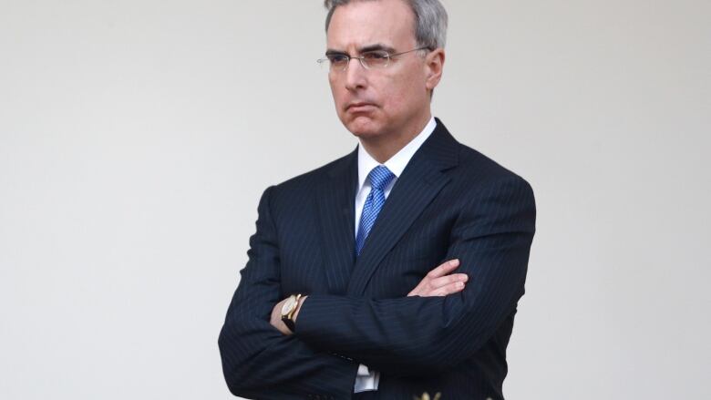 A serious looking individual wearing glasses and a blue tie, with arms crossed.