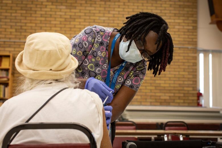 A nurse wearing a blue medical mask and latex gloves administers a vaccine to the right arm of a woman with grey hair, wearing a hat.