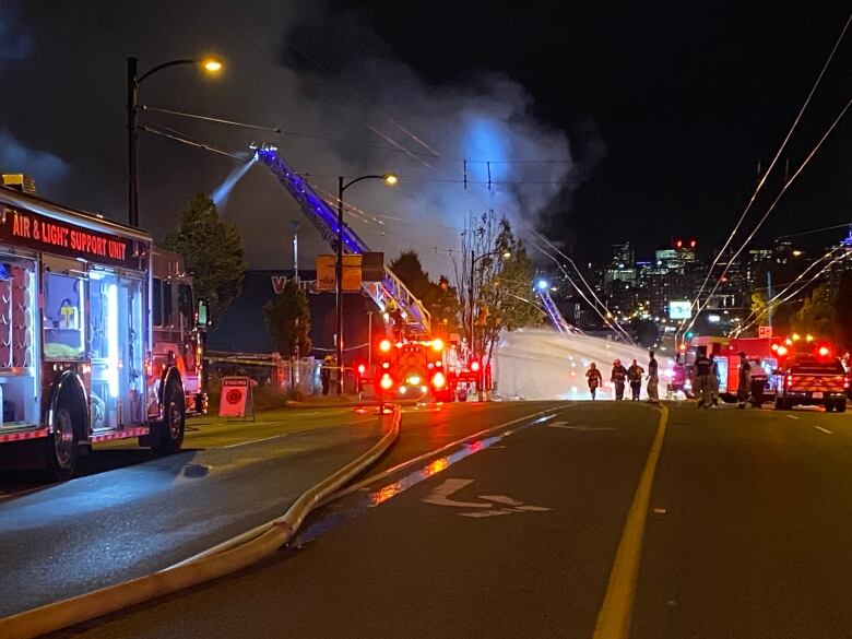 Three fire engines are seen with flashing lights, smoke is visible in the background on a closed street.