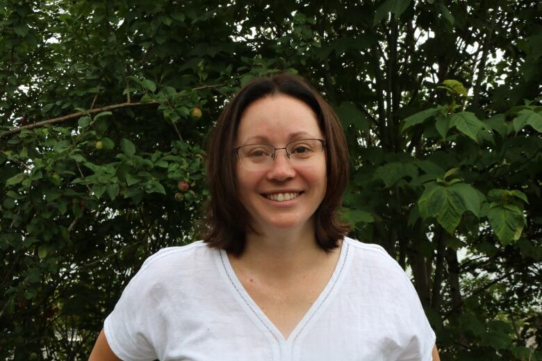 A woman wearing glasses and a white T-shirt smiles at the camera. Green leaves of an apple tree are seen behind her.