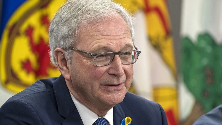 a closeup of a man wearing a jacket and tie, and sitting at a table in front of the New Brunswick flag