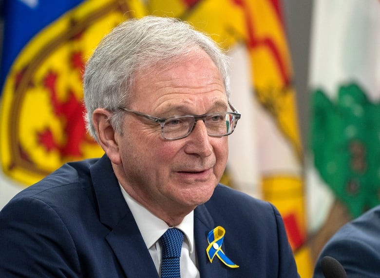 a closeup of a man wearing a jacket and tie, and sitting at a table in front of the New Brunswick flag