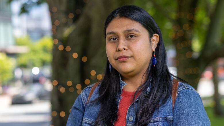 Stacey Gomez has long, black hair and is wearing a blue denim jacket. She is looking at the camera with lights and trees in the background.