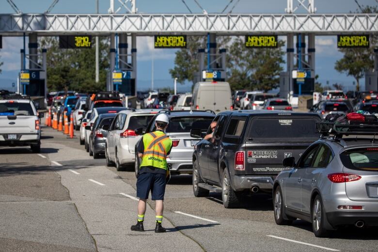 A person in a high-vis jacket stands on the road among dozens of lined-up cars.