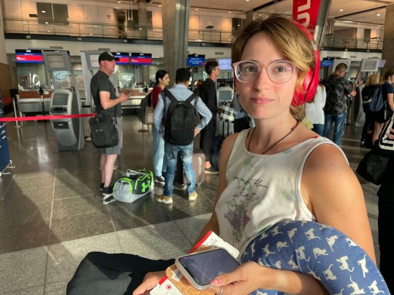 A woman sits in an airport. She is wearing headphones and holding her phone.