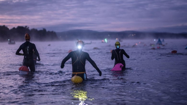 People in wetsuits, wearing headlamps, head into a cold, dark lake before dawn.