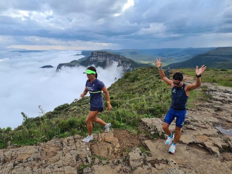 A man and woman run along rugged terrain at the edge of a cliff, above the clouds.