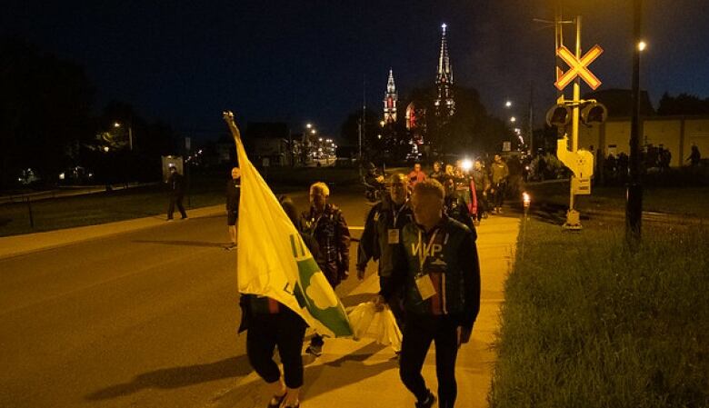 A long line of people walk in the dark, a church lit up in the background.