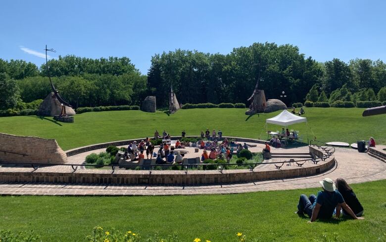 Two people sit in the grass looking on at a larger group of people sitting in a circle.