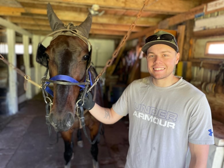 Young man, smiling, stands next to brown horse in a stable.