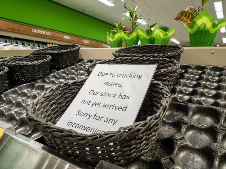 An empty basket among empty shelves. A sign reads 'due to trucking issues, our stock has not yet arrived. Sorry for any inconvenience.'