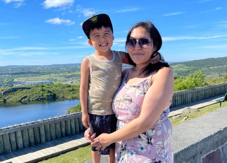 A person wearing a floral dress holds a small child as he stands on a stone wall on Signal Hill on a sunny day.