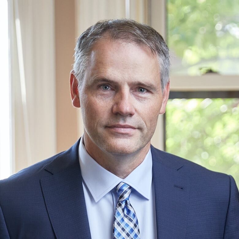 A man with grey hair poses for a head shot in a blue suit and tie.