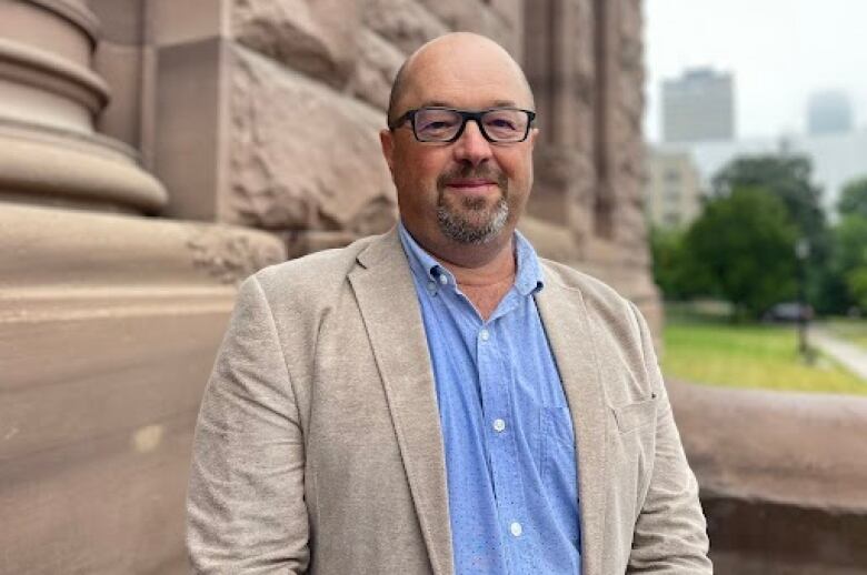 A man wearing glasses stands outside in front of a large stone building.