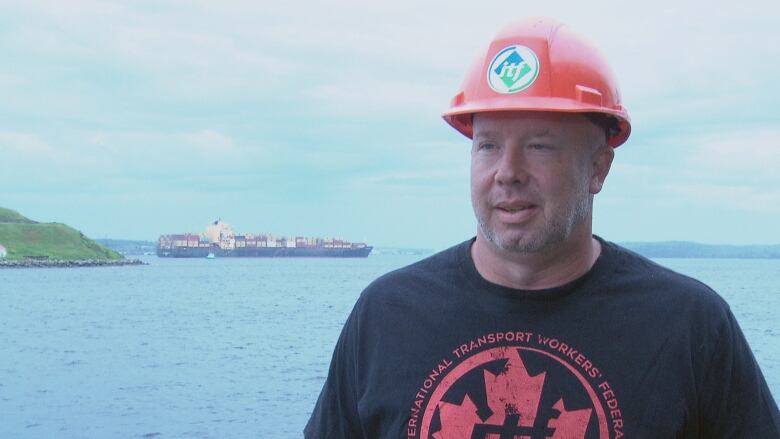 Karl Risser stands in front of the Halifax Harbour. George's Island and a detained cargo ship can be seen in the background.