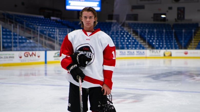 A teenage boy dressed in hockey gear holds a hockey stick and helmet on the ice. 