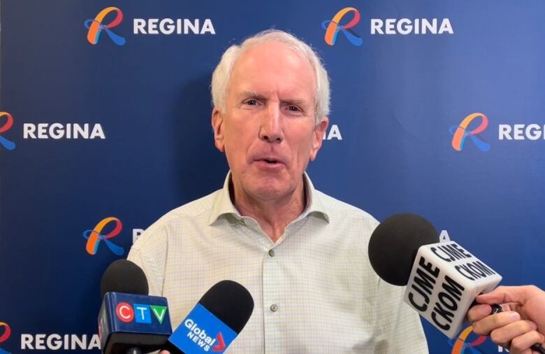 A man in a light button-up shirt and grey hair speaks into microphones.