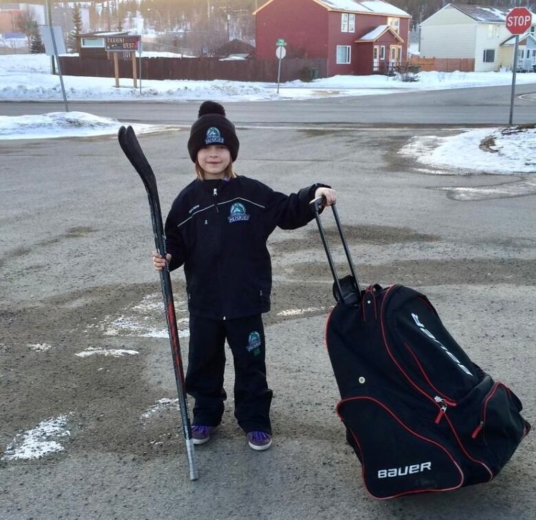 A young boy wearing a toque stands outside holding a hockey stick and pulling a large hockey equipment bag. 