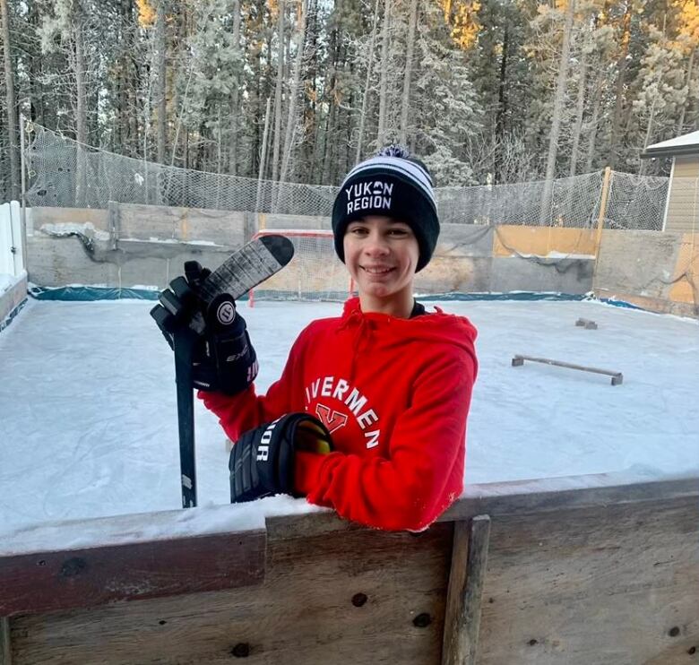 A smiling young boy holding a hockey stick leans over the boards of a backyard ice rink. 