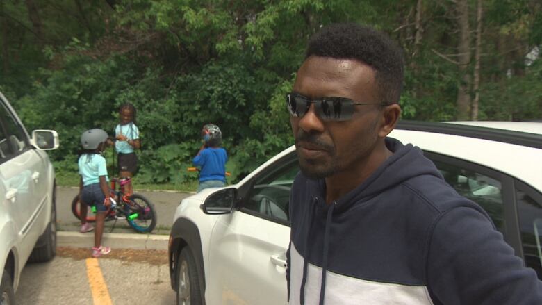 A man in sunglasses stands in the foreground as three young kids, some with bicycles, stand behind him.