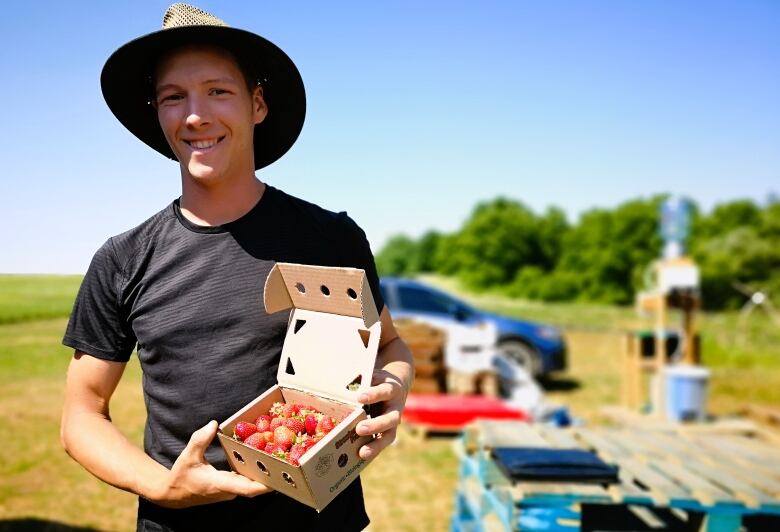 A man wearing a hat stands in a field holding a box of strawberries.