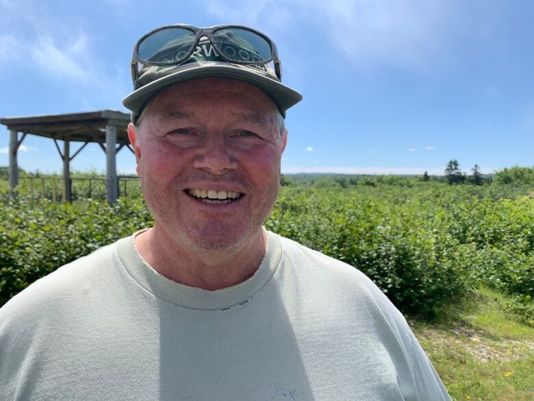 A man stands in a field, smiling, wearing a baseball hat and sunglasses on a sunny day.