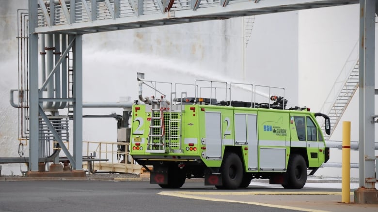 A green box-shaped fire truck sprays foam from a nozzle mounted on top in amongst large fuels storage tanks.