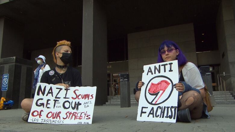 Two young women sit in front of the court. One sign reads, 'Nazi scum, get off of our streets.'