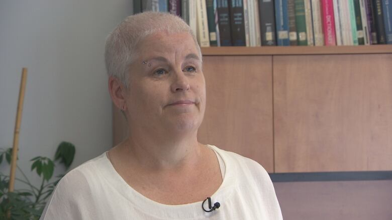 A woman with buzzed white hair and an eyebrow piercing stands in front of a bookcase.