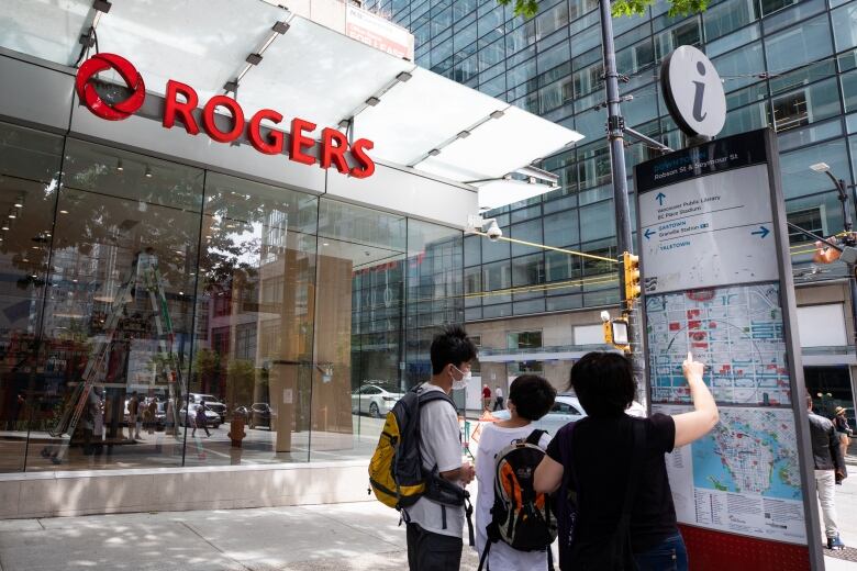 A group of people point at a street map in the shadow of a Rogers store on a street.