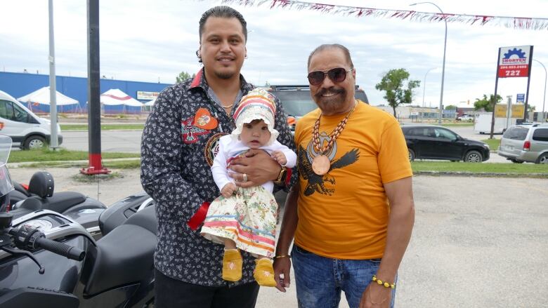 Two men, one holding a small child, stand smiling in a parking lot next to some motorcycles. The child is wearing a ribbon-decorated bonnet and ribbon skirt.