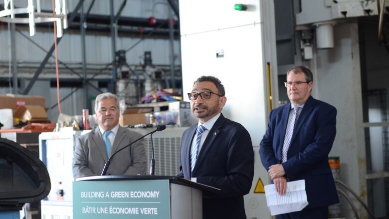 Three man stand in an industrial building. One is speaking into a microphone. 