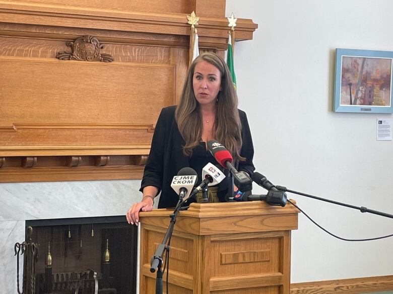 A woman stands at a podium with several media-branded microphones in front of her.