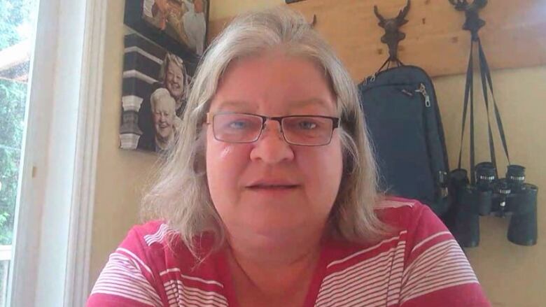 A close-up of a woman with shoulder-length, grey hair and glasses, wearing a fuchsia and white striped shirt.