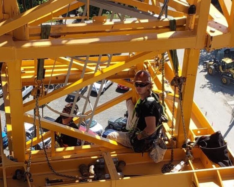 A man wearing a hard hat and helmet is shown sitting in a structure composed of yellow girders that are part of a crane assembly.