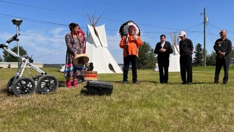 A chief speaks near a piece of equipment in front of two teepees as four other people stand nearby.