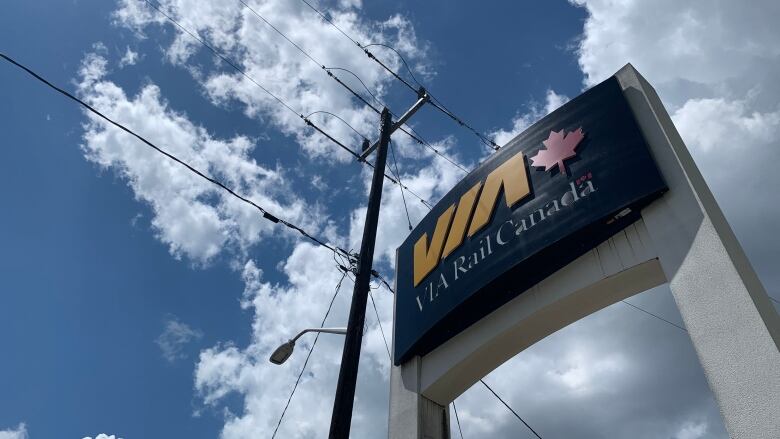 Via rail sign is shown from below with a blue, cloudy sky and utility lines behind it.